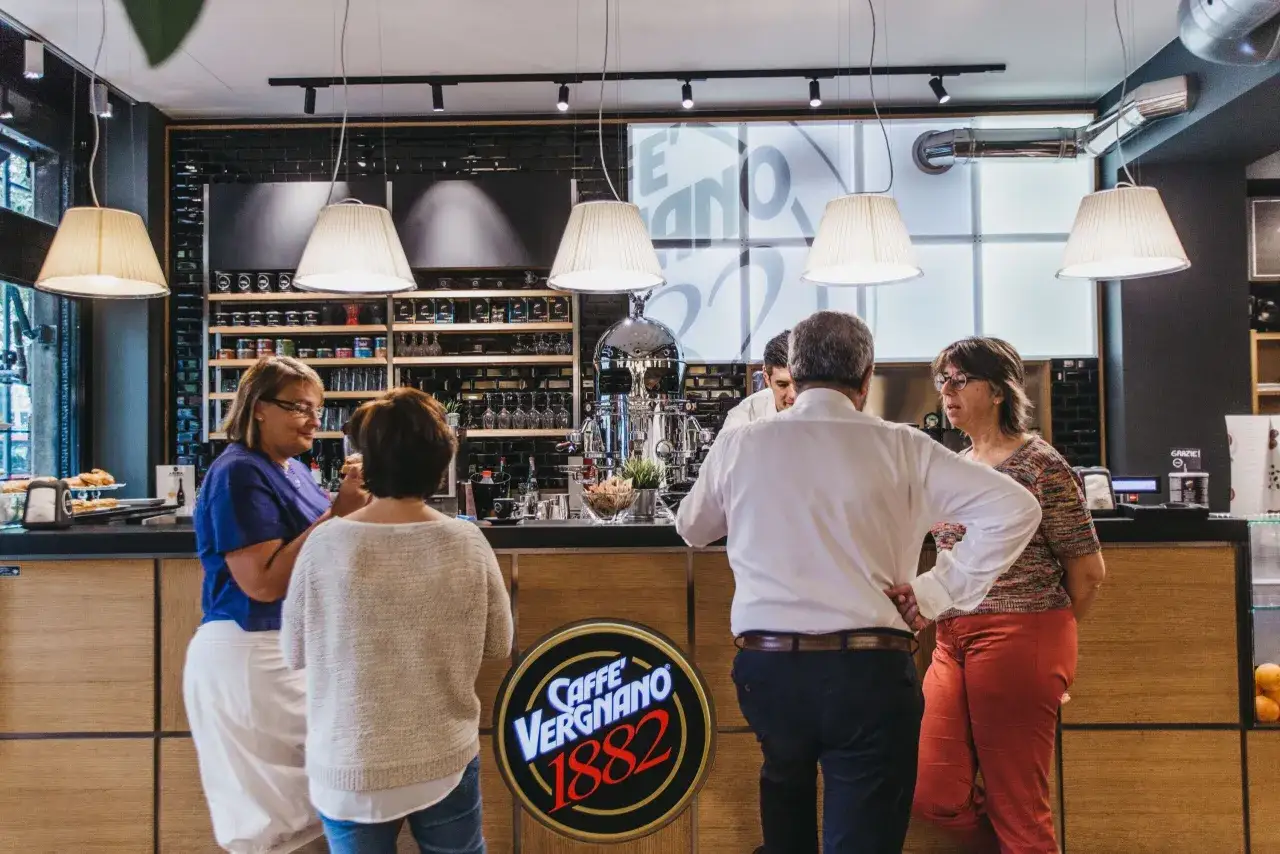 Persone al bancone di un caffè, con un barista che serve. Il logo 