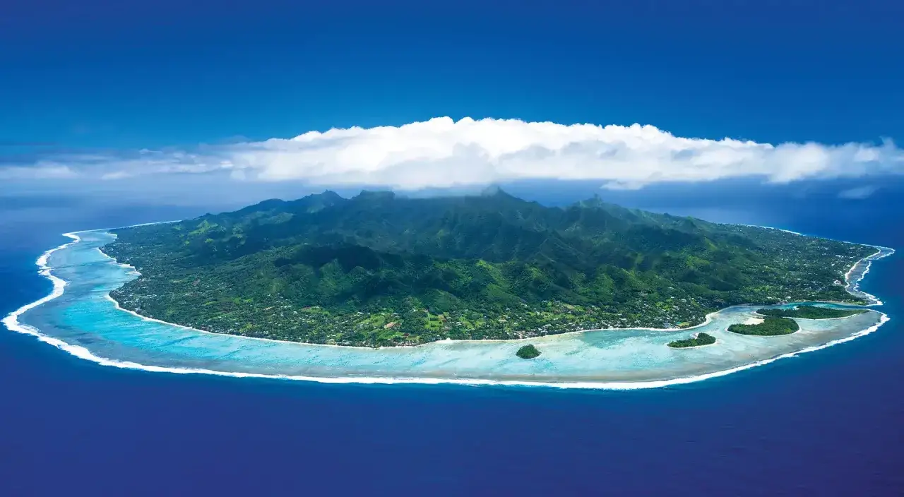 Lush green mountains rise from a tropical island in the Cook Islands, surrounded by turquoise lagoons and the deep blue ocean.