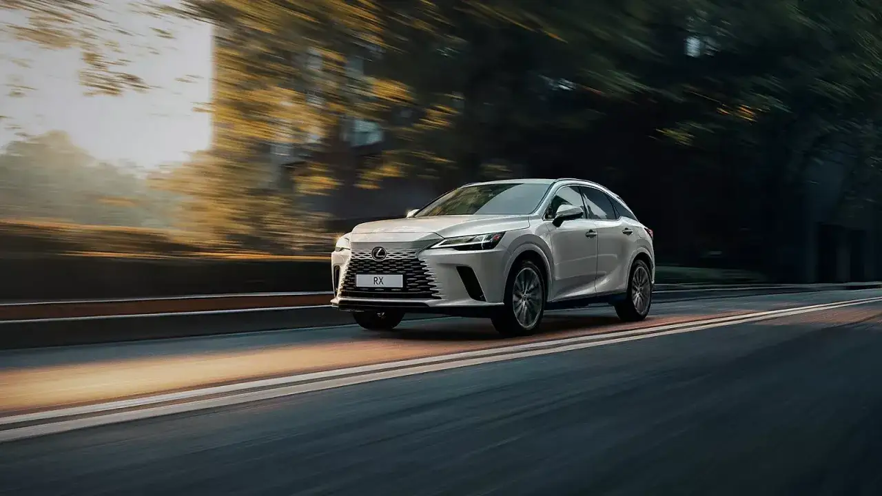 Biały Lexus RX 350h pędzi po drodze, otoczony rozmazanymi drzewami i światłem zachodzącego słońca.