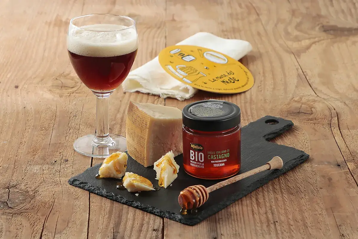 Un bicchiere di birra ambrata, formaggio stagionato e pezzetti di formaggio conditi con miele di castagno. Un perfetto abbinamento cibo birra per un momento di gusto.