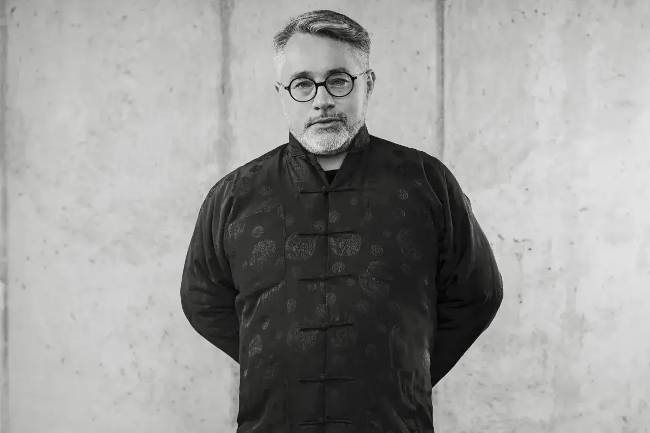 Mężczyzna w okularach i czarnym, wzorzystym **robak wruhme** stoi na tle betonu.