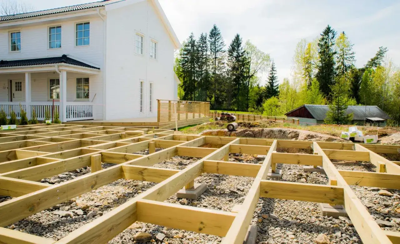 Grundläggande stomme för altanen är på plats, redo att skruva trall. Ett vitt hus och grönskande träd i bakgrunden.