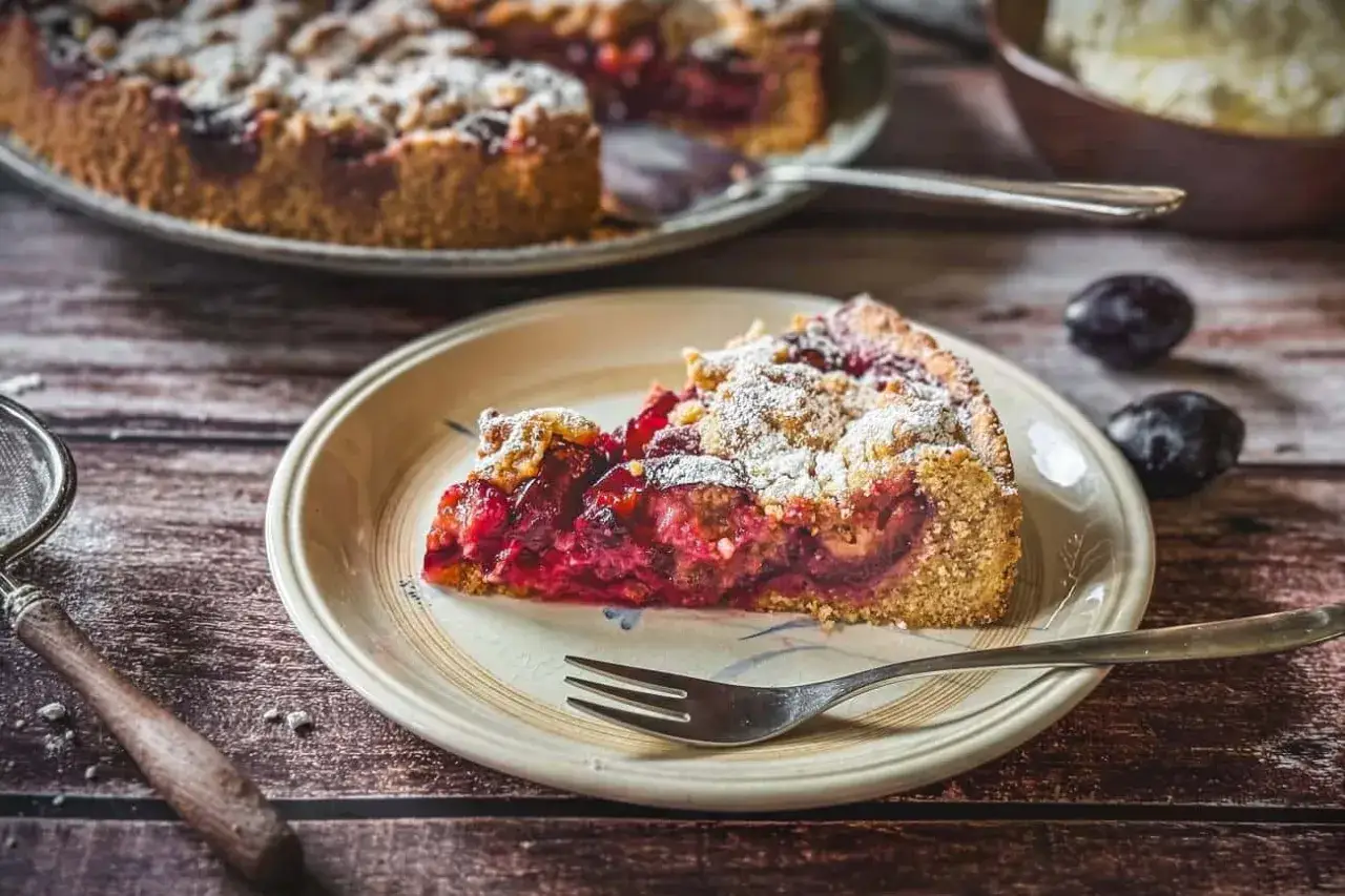 Ein saftiges St&uuml;ck Zwetschgenkuchen mit Dinkelmehl vom Blech, best&auml;ubt mit Puderzucker. Daneben liegen frische Zwetschgen.
