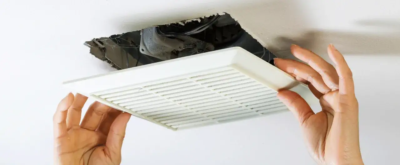 Mains d'une personne retirant la grille d'un ventilateur de salle de bain, révélant la poussière et les toiles d'araignées à l'intérieur.