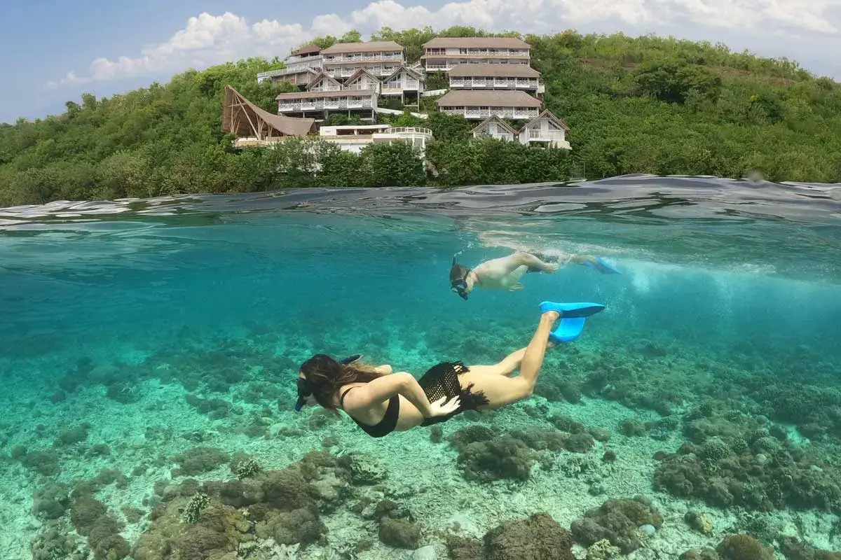 Idealne wakacje na Bali: para nurkuje w krystalicznie czystej wodzie przy rafie koralowej, w tle hotel na wzgórzu.