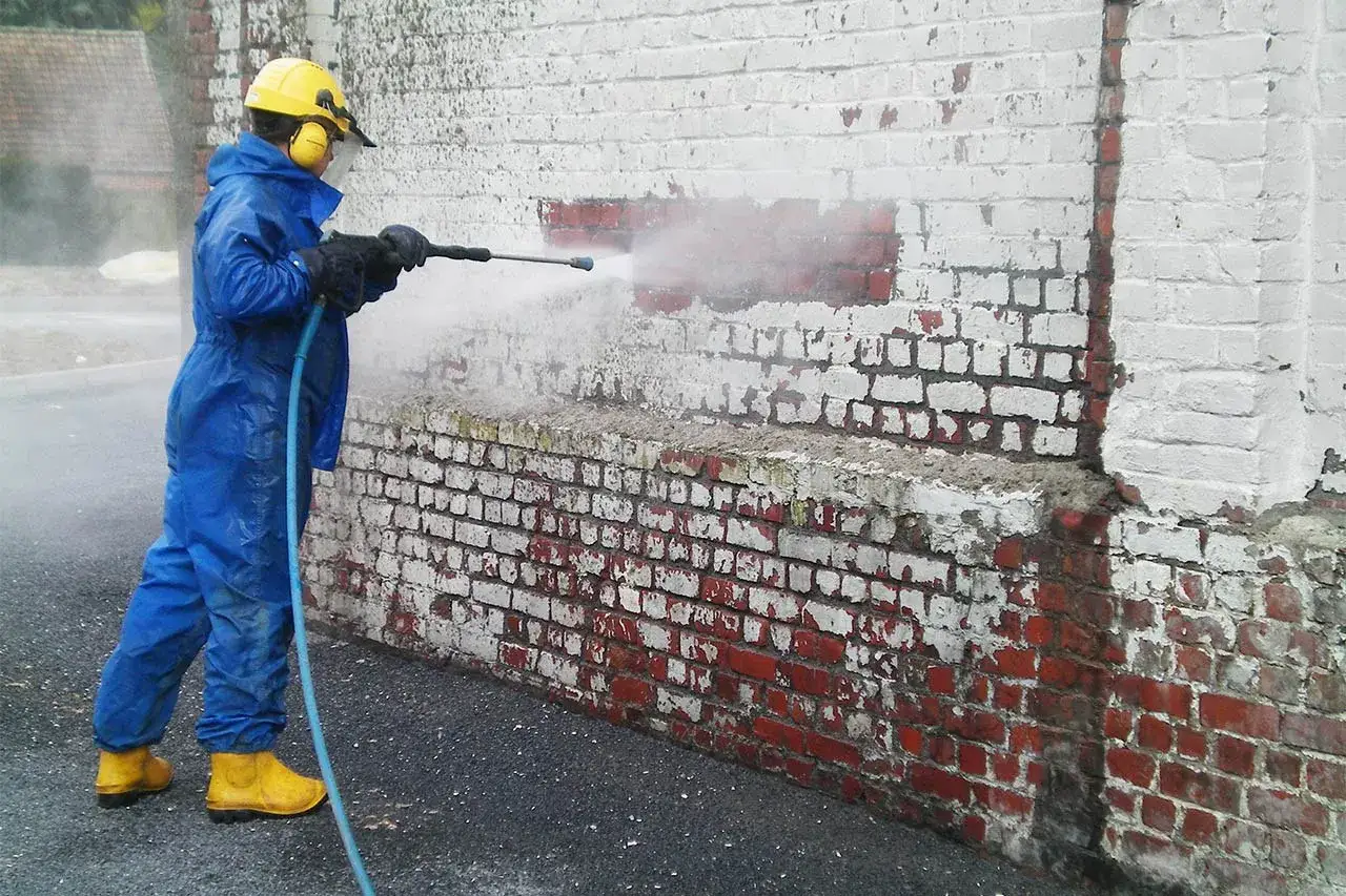 Travailleur en combinaison bleue et casque jaune utilise un nettoyeur haute pression pour enlever peinture sur brique.