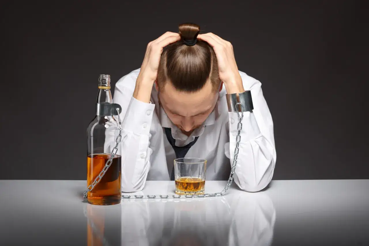 Mężczyzna w kajdanach, przykuty do butelki i szklanki z alkoholem, symbolizuje nadużywanie alkoholu co to znaczy: zniewolenie i rozpacz.