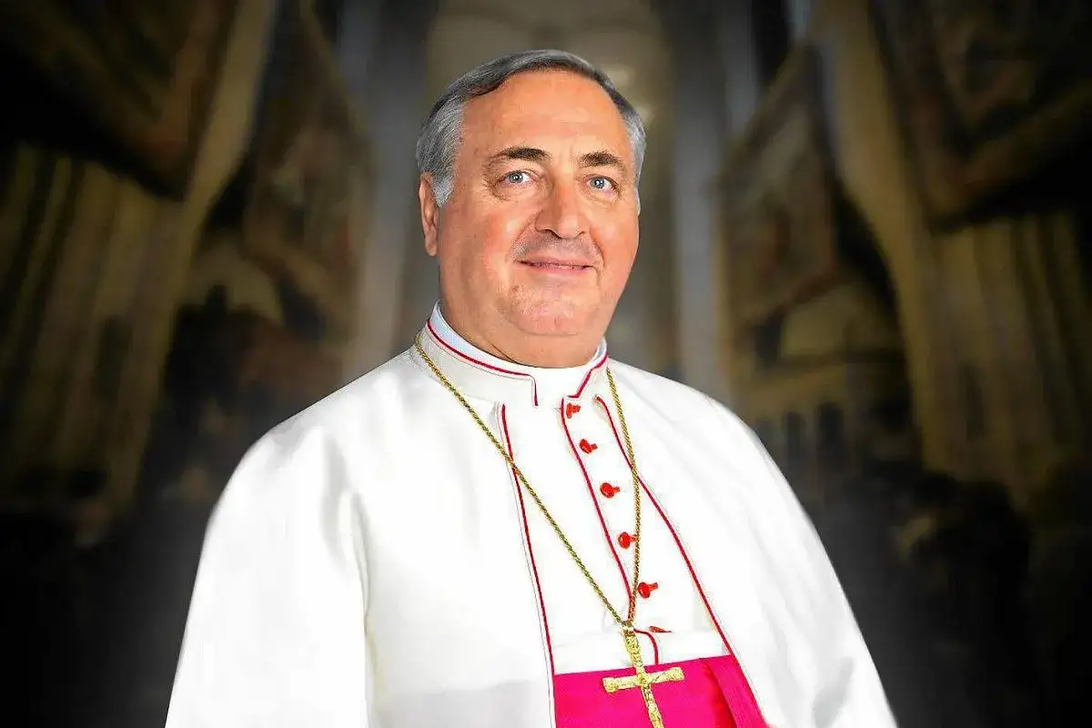 Uśmiechnięty nuncjusz apostolski w białej szacie z czerwonymi zdobieniami i złotym krzyżem na szyi.