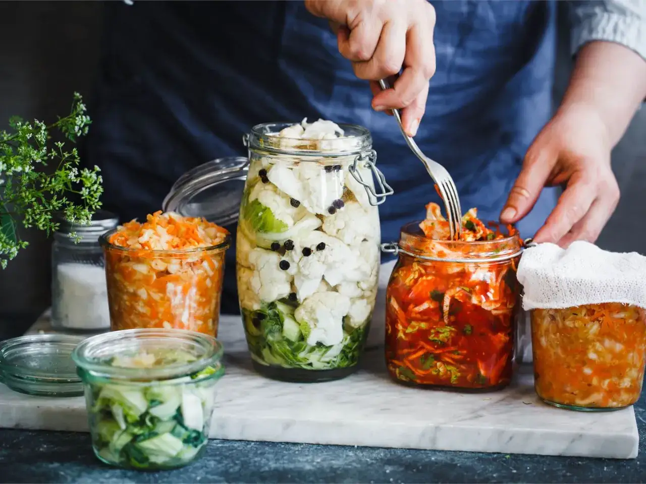 H&auml;r &auml;r flera burkar med fermenterad mat, inklusive blomk&aring;l, mor&ouml;tter och k&aring;l. En person anv&auml;nder en gaffel f&ouml;r att plocka ur kimchi.