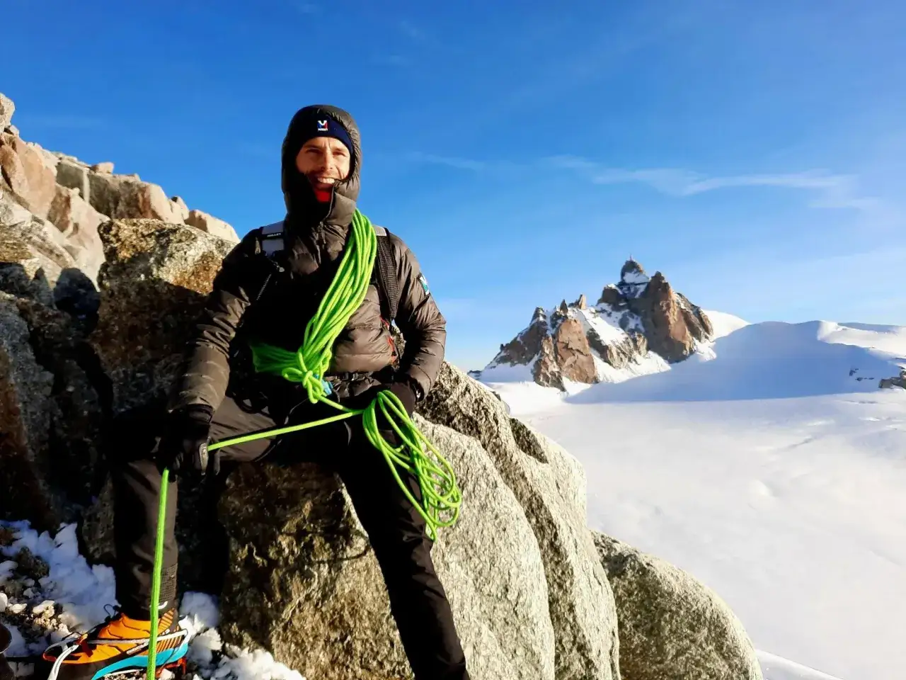 Un alpiniste souriant, corde verte enroulée, sur un rocher enneigé. Le décor parfait pour un podcast alpinisme, avec des sommets enneigés en arrière-plan.