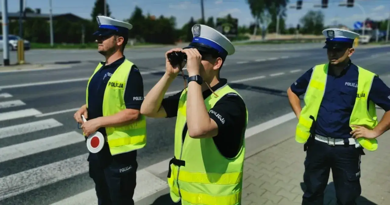 Trzech policjantów w kamizelkach odblaskowych na przejściu dla pieszych. Jeden obserwuje przez lornetkę, drugi trzyma lizak, trzeci stoi z rękami na biodrach. Czyżby szukali dodatkowej masy w samochodzie?
