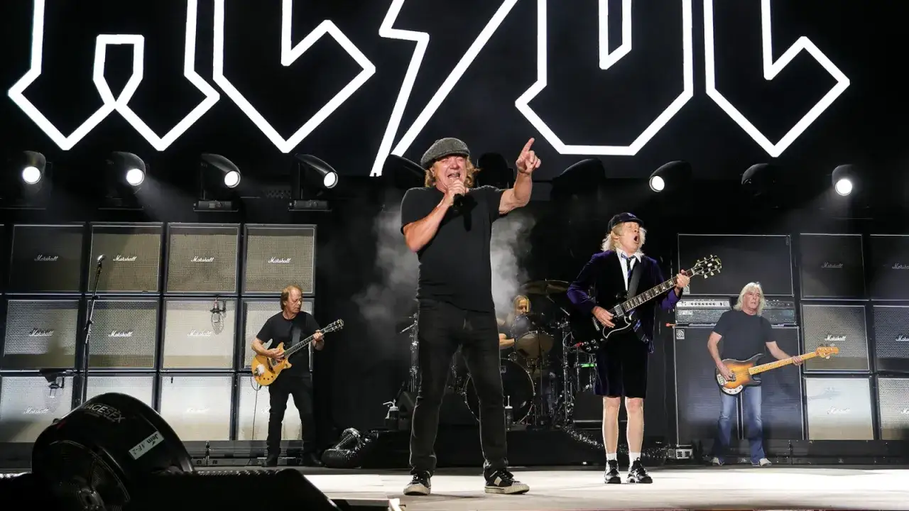 Zespół AC/DC w pełnym składzie na scenie, z charakterystycznym logo w tle. Muzycy grają na gitarach i śpiewają, tworząc energetyczny koncert.