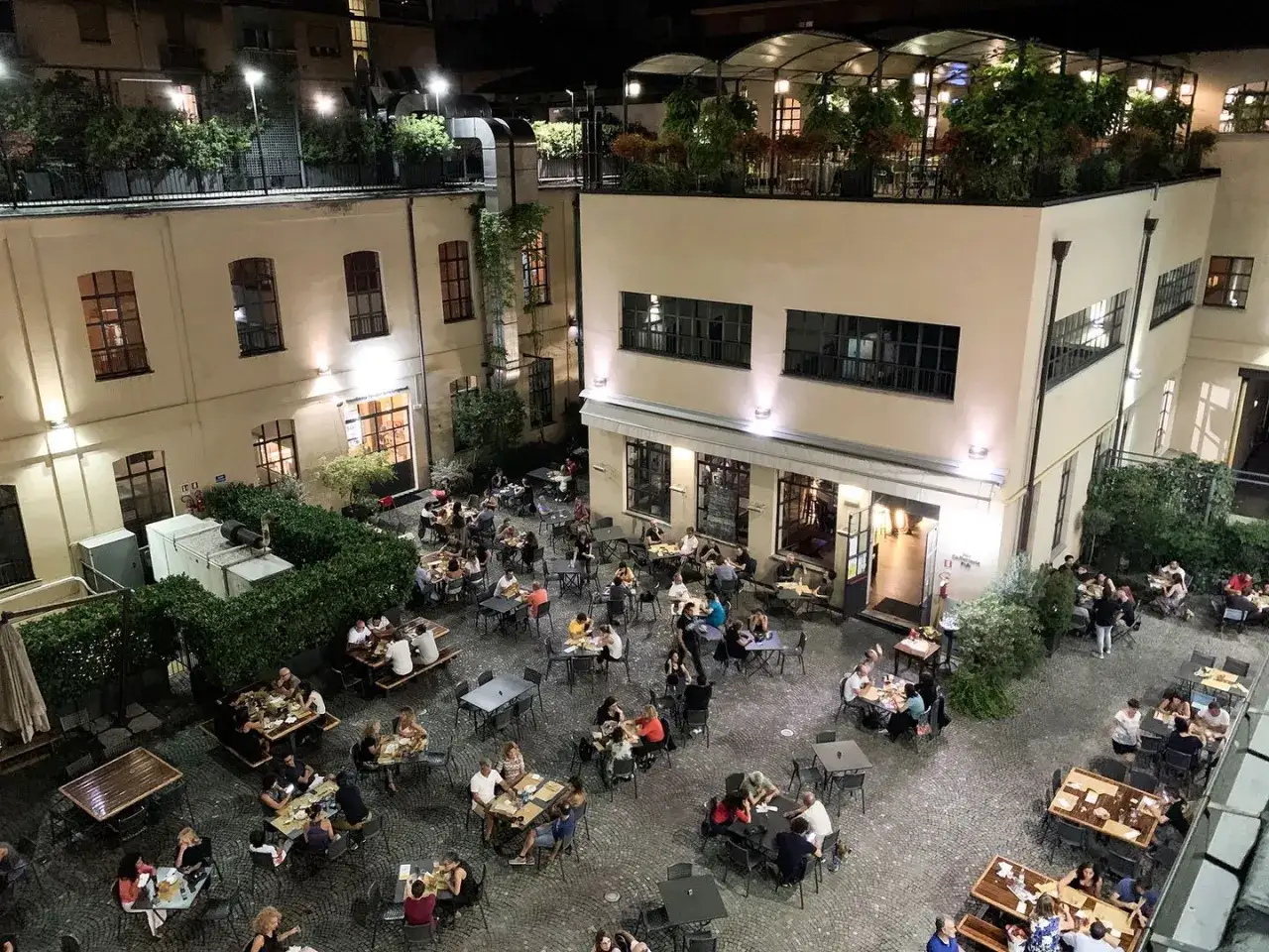 Serata vivace al birrificio La Piazza, con tavoli all'aperto gremiti di persone che gustano cibo e bevande sotto le luci soffuse.