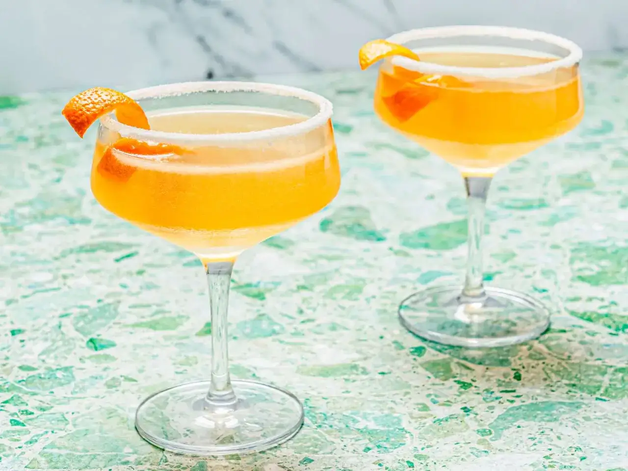Zwei Sidecar Cocktails mit Zuckerrand und Orangenzeste auf einem gr&uuml;nen Marmortisch.