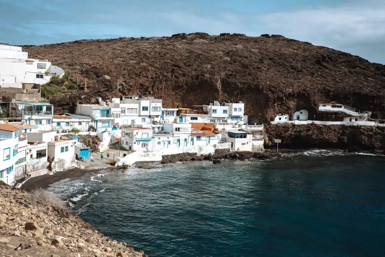 Ein malerisches Dorf mit wei&szlig;en H&auml;usern und blauen Akzenten schmiegt sich an eine dunkle Felswand. Ein versteckter Ort, einer der Gran Canaria Geheimtipps.