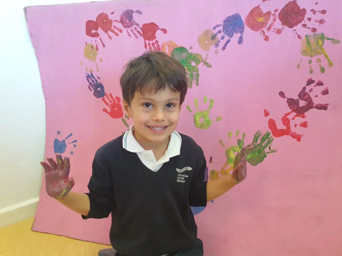 Ein Junge mit bemalten Händen lacht vor einer rosa Wand mit einem Herz aus bunten Handabdrücken. Perfekt für Basteln zum Thema Freundschaft Grundschule.