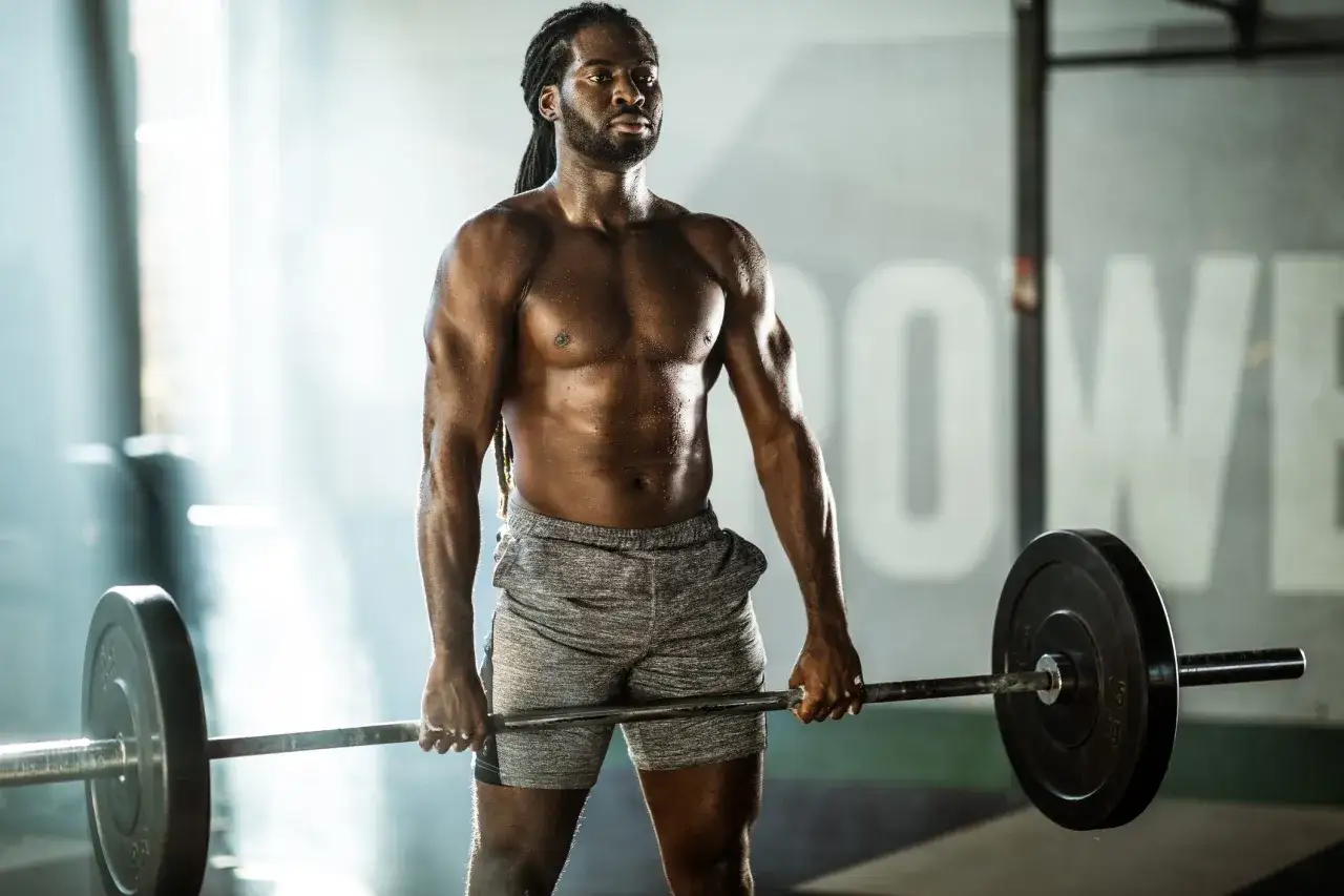 Ein Mann mit Dreadlocks und muskulösem Körper hält eine Langhantel. Sein intensives **bodybuilding training** zeigt Schweißperlen auf seiner Haut.