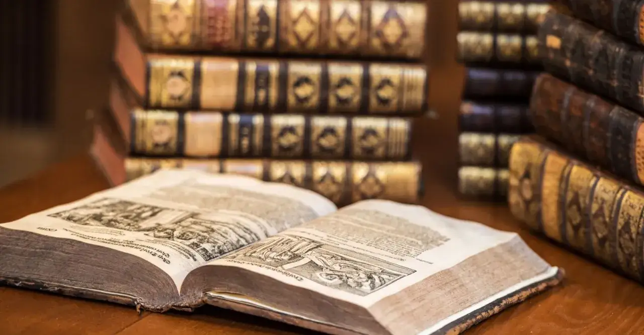 Antichi libri rilegati in pelle, con decorazioni dorate, disposti su un tavolo di legno. Alcuni sono impilati, uno è aperto mostrando illustrazioni e testo antico. Tesori di sapere, questi libri costosi raccontano storie di epoche passate.