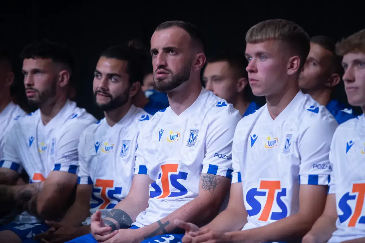 Młodzi piłkarze Lecha Poznań w białych strojach, siedzą obok siebie. Kadra gotowa do walki.
