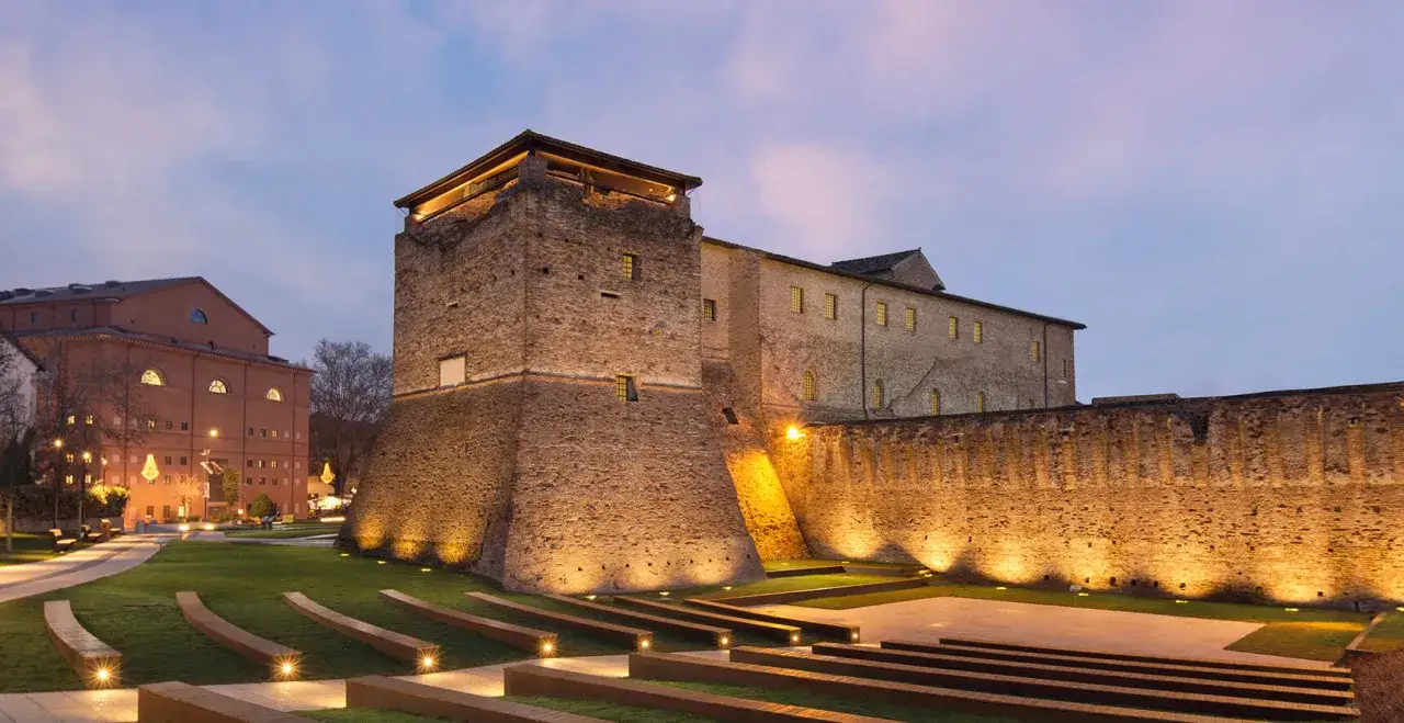 La Rocca Malatestiana di Rimini illuminata di sera, con un parco moderno e un edificio in mattoni rossi sullo sfondo.