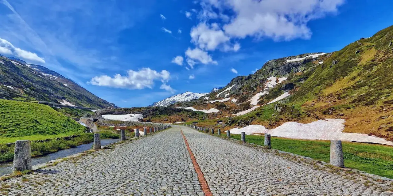 Strada acciottolata sul Grimsel Pass, con montagne verdi e nevicate residue sotto un cielo azzurro.