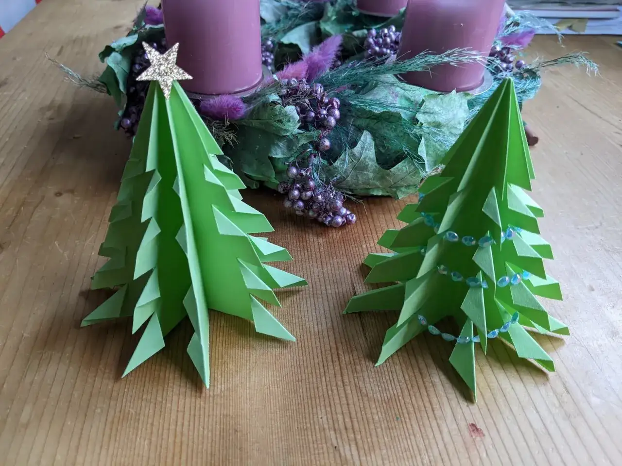 Zwei grüne **origami tannenbaum flach** stehen vor einem Adventskranz mit lila Kerzen. Einer ist mit einer goldenen Sternspitze verziert.