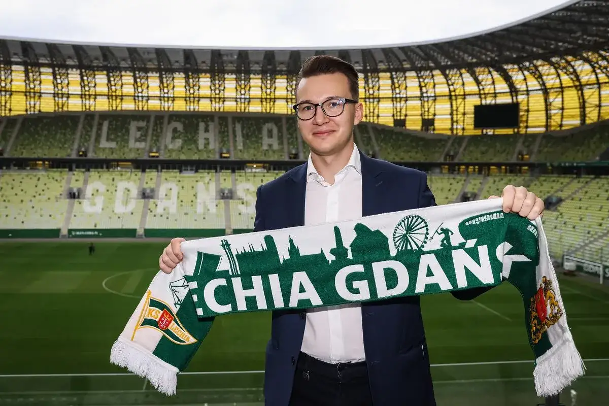 Mężczyzna w garniturze prezentuje szalik Lechii Gdańsk na tle stadionu. Zapowiada nowe lechia transfery.