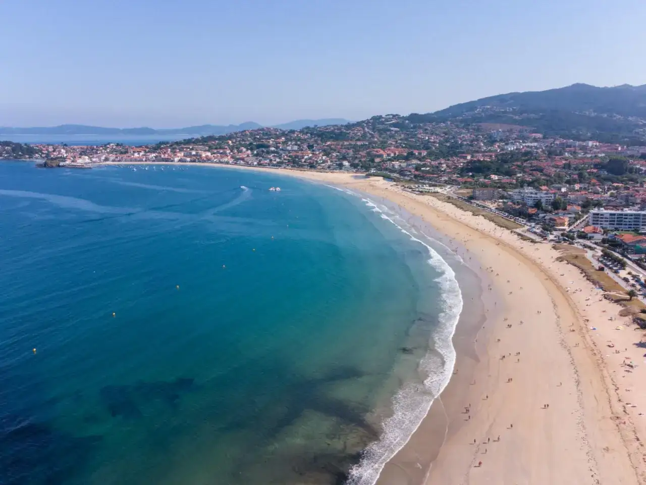 Vista aérea de la playa América, con aguas turquesas, arena dorada y un pueblo costero al fondo.