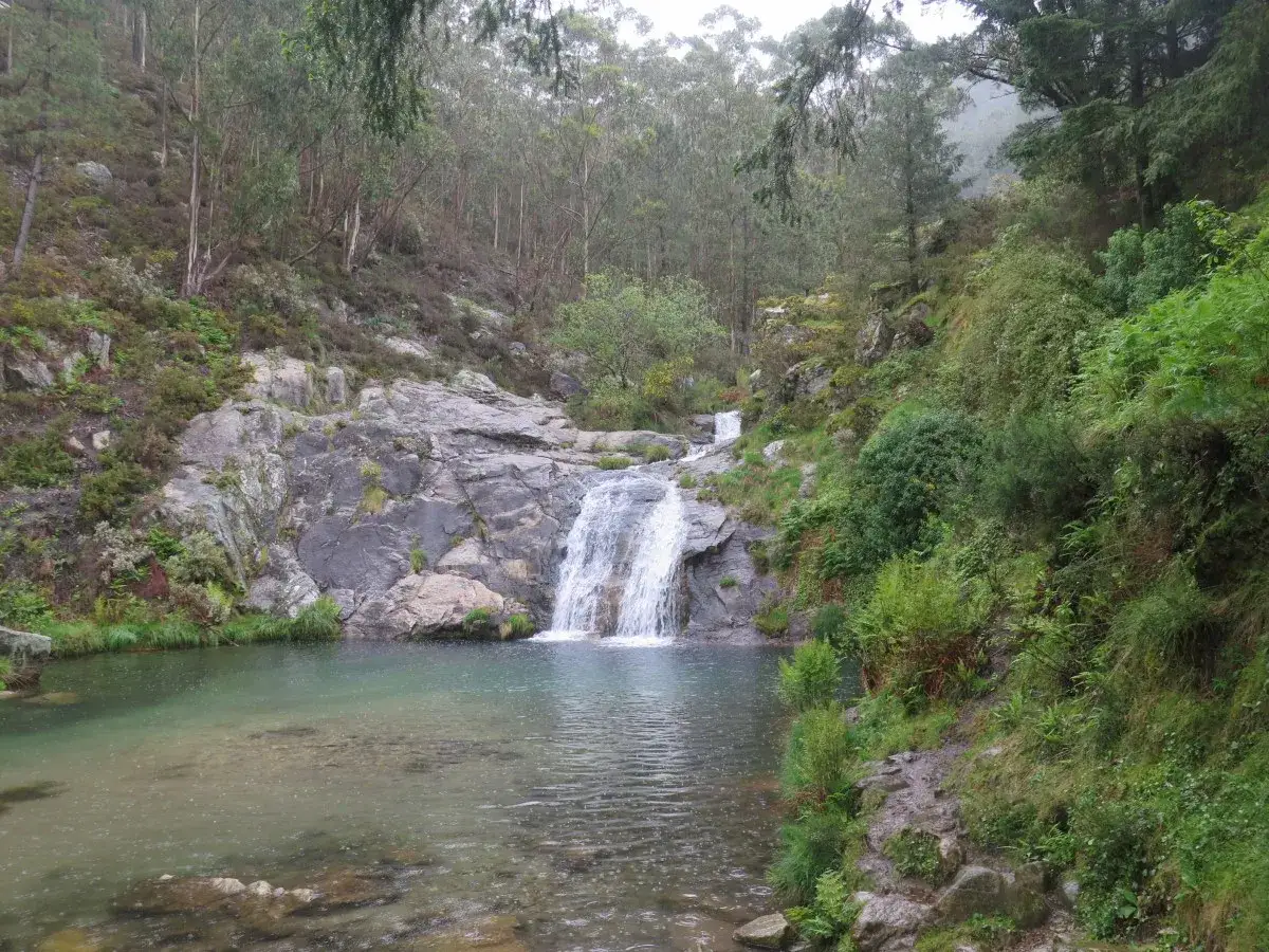Cascada y poza natural en las Pozas de Mougas, rodeada de vegetación frondosa y rocas.