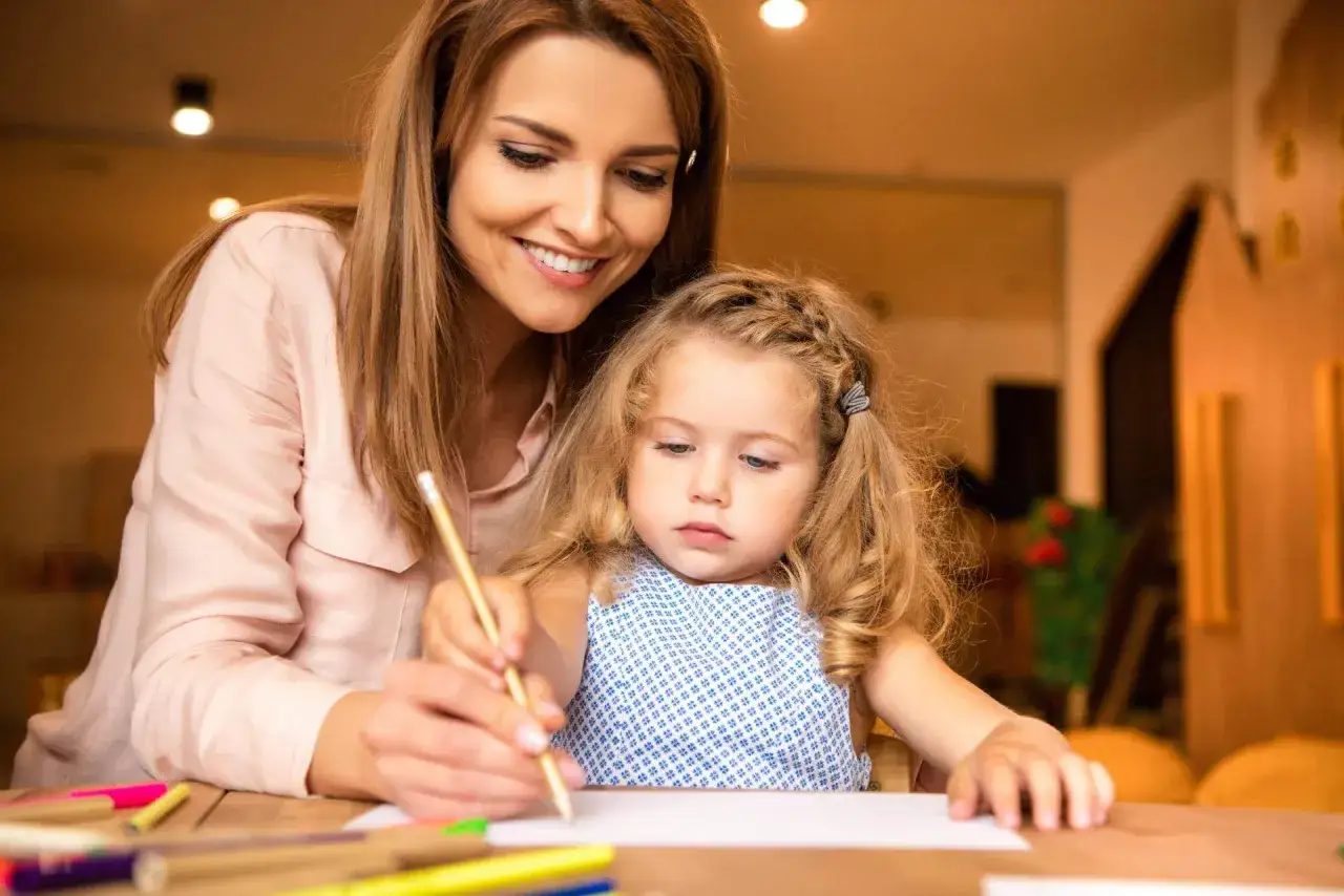 Donna sorride mentre guida la mano di una bambina che disegna. Un'immagine che evoca la gioia di chi cerca offerte lavoro educatore.