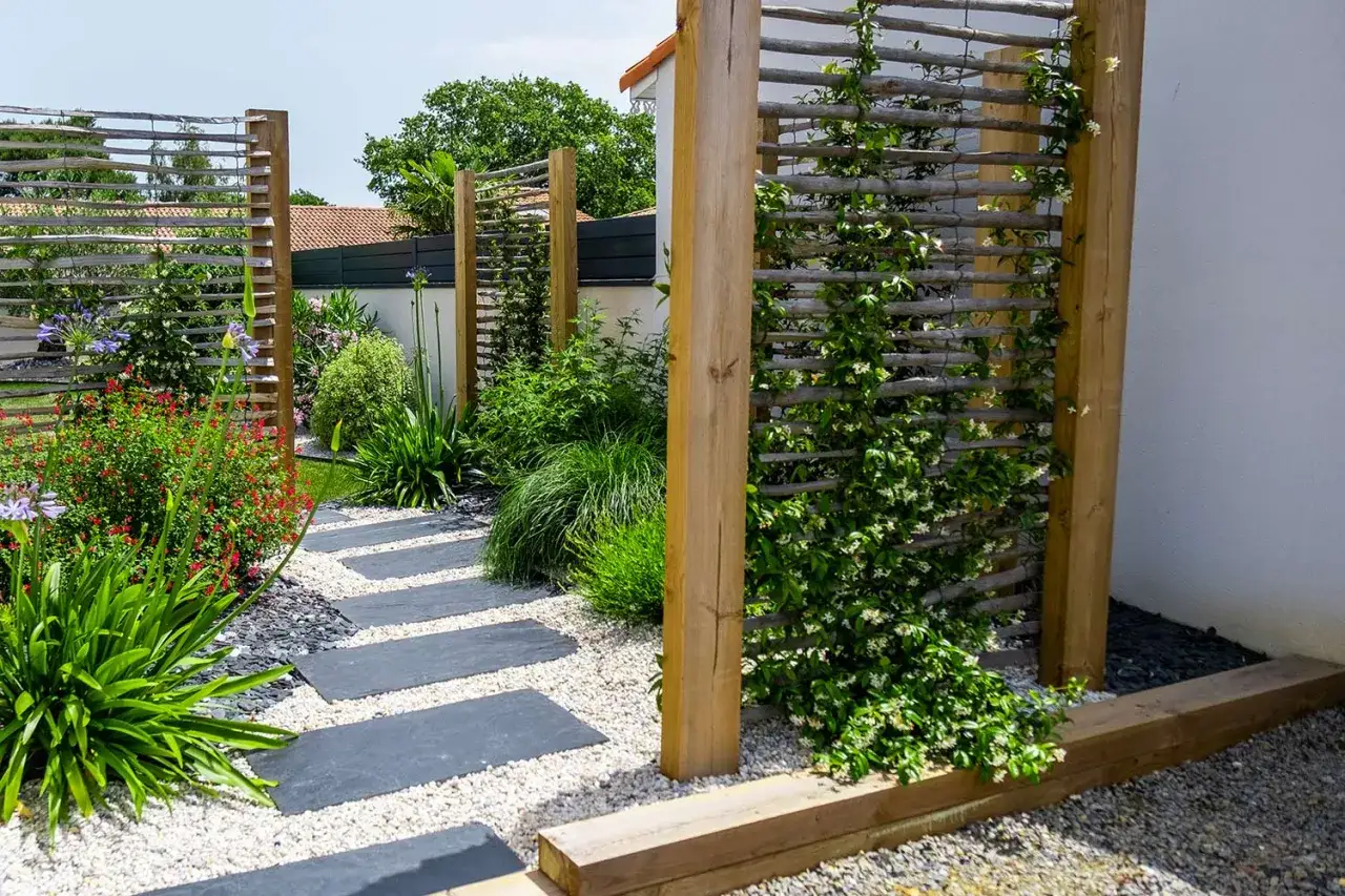 Un chemin de dalles grises serpente &agrave; travers un jardin luxuriant. Des treillages en bois servent de **plante brise vue**, orn&eacute;s de v&eacute;g&eacute;tation grimpante et de fleurs.