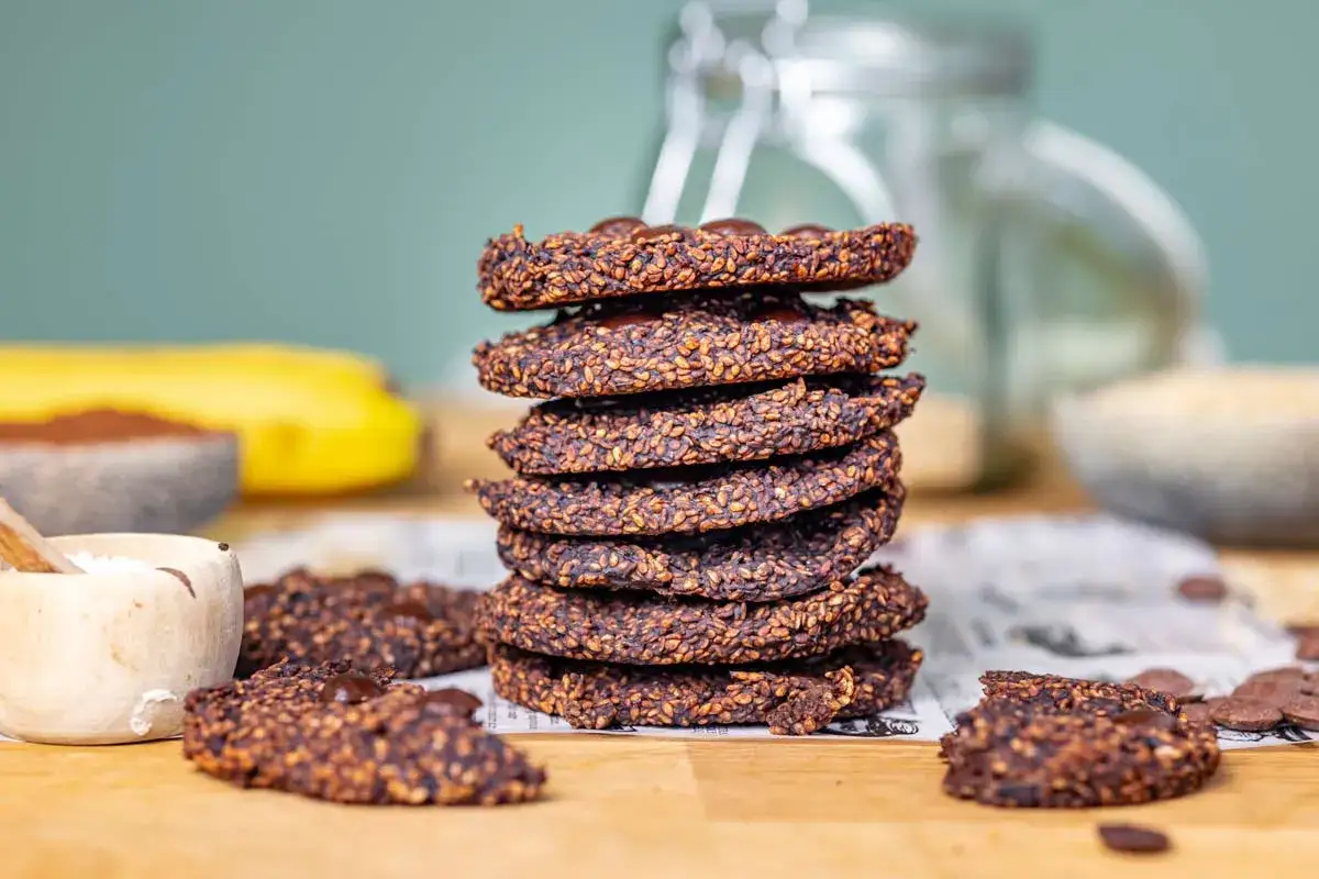 Gestapelte Bananen-Cookies mit Sesam und Schokostückchen, daneben eine Banane und Kakaopulver.