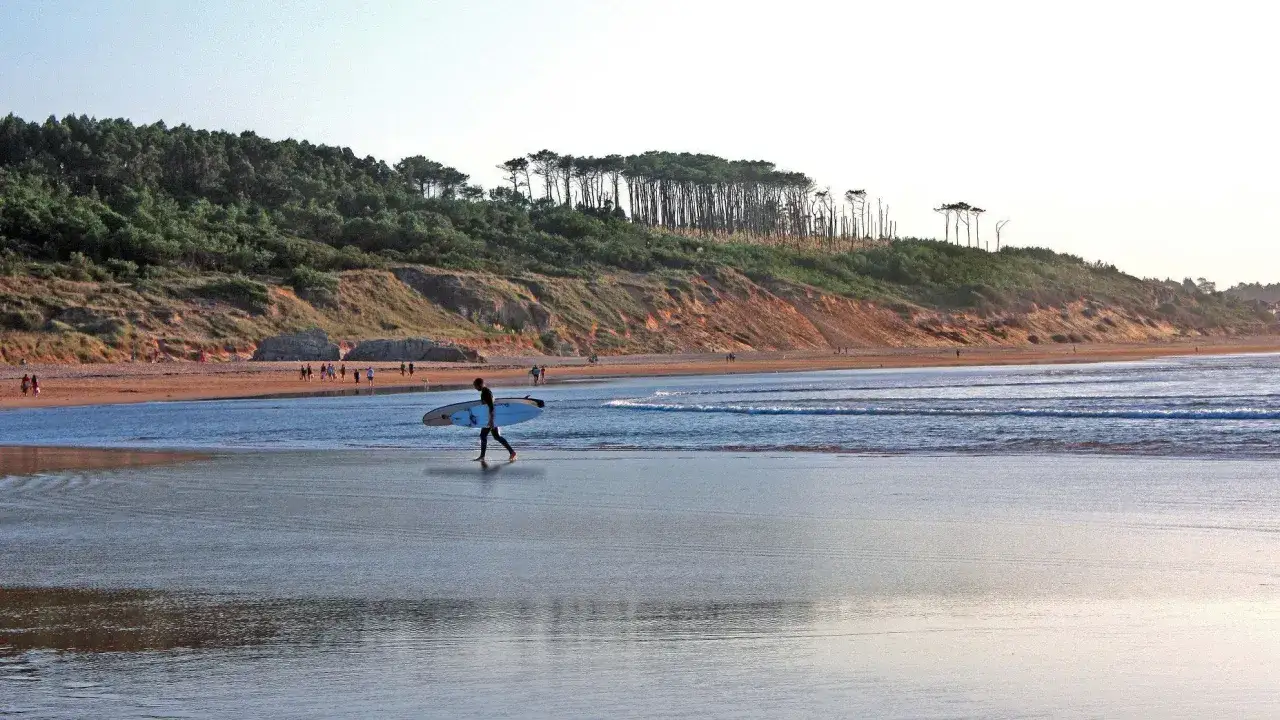 Un surfista camina por la playa de Somo con su tabla bajo el brazo, mientras la gente pasea por la orilla y las olas rompen suavemente.