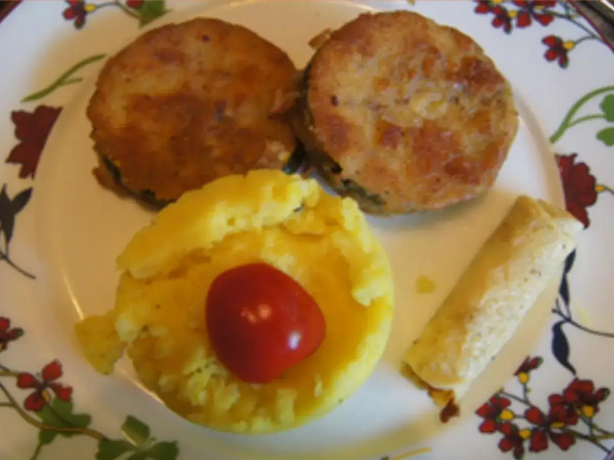 Zucchini-Schnitzel, Kartoffelbrei mit Kirschtomate und eine kleine Roulade auf einem Teller mit Blumenmuster.