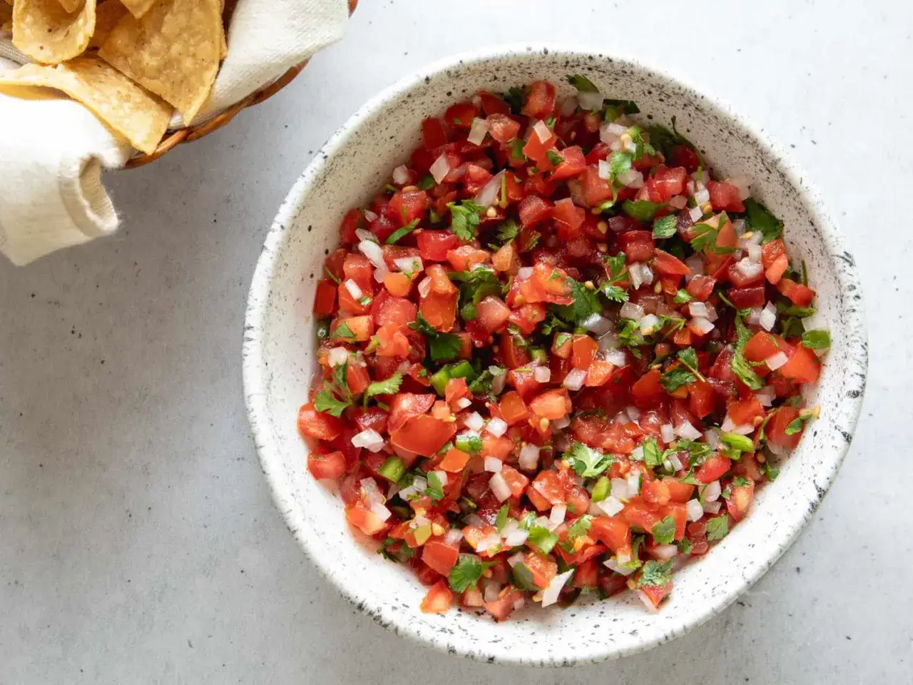 En skål med färsk pico de gallo, full av hackade tomater, lök och koriander. Bredvid syns en korg med tortillachips.