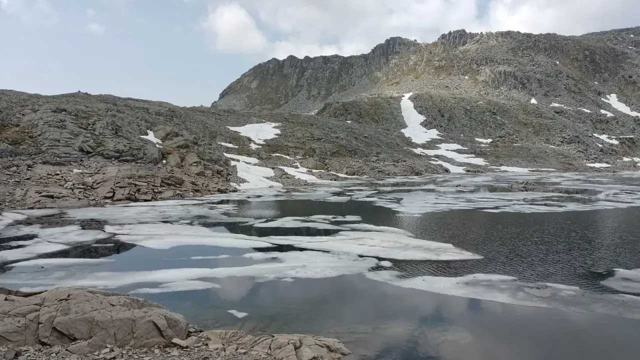 Il lago della Vacca, con rocce scure e ghiaccio che galleggia sull'acqua, sotto un cielo nuvoloso e montagne imponenti.