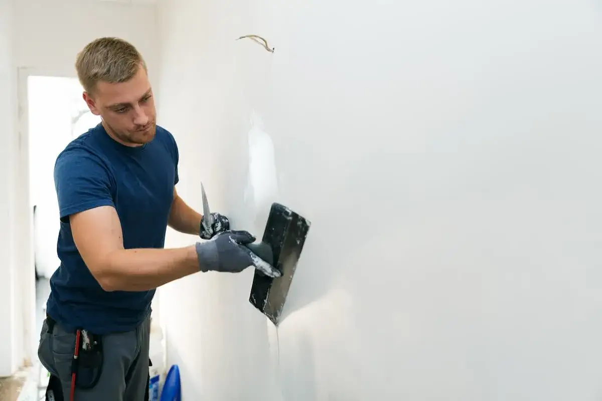 Mann mit Spachtel und Kelle bearbeitet eine Wand, um alte Latexfarbe zu entfernen und die Fl&auml;che f&uuml;r neue Farbe vorzubereiten.