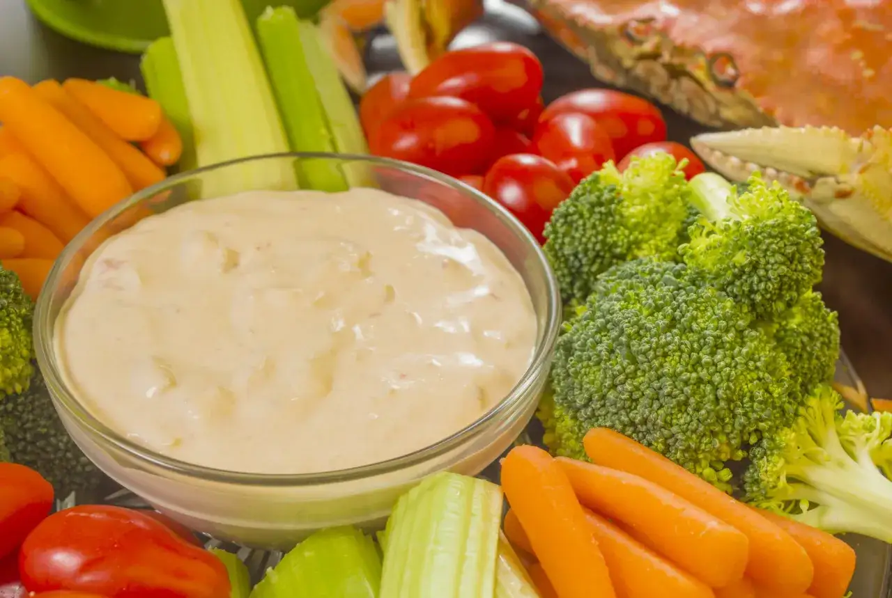 Un bol de sauce crudités crème fraîche entouré de carottes, céleri, brocolis et tomates. Un crabe cuit est visible en arrière-plan.