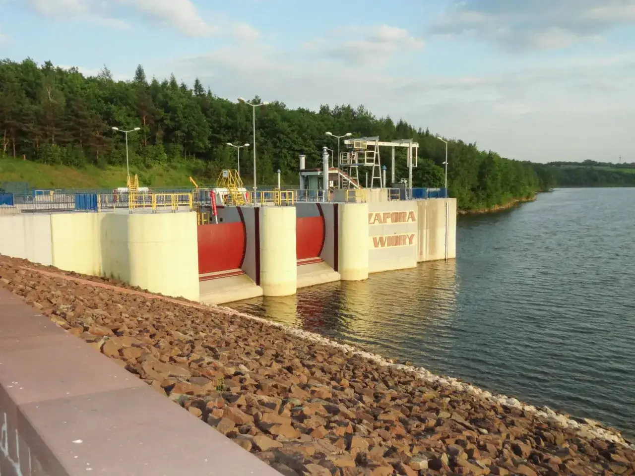 Zapora Wióry, potężna konstrukcja na tle zielonych drzew, chroni przed powodzią. Woda spokojnie spływa po kamiennym brzegu.