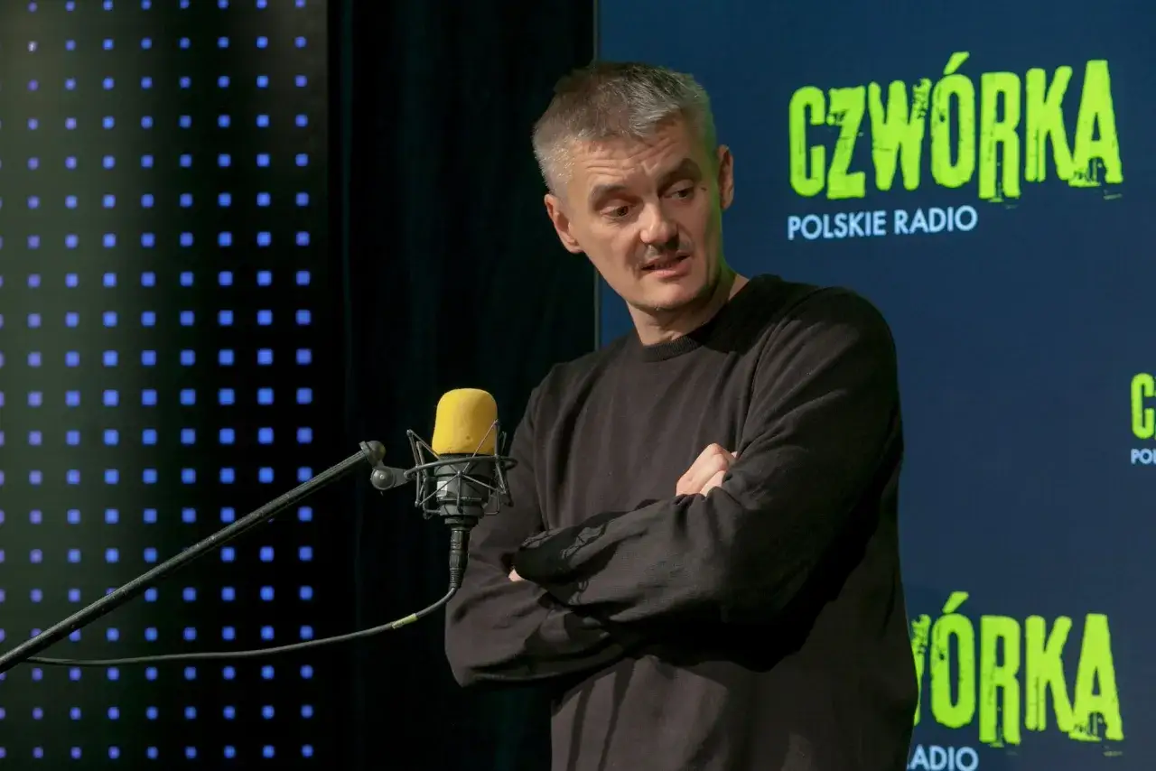 Jacek Sienkiewicz w studiu radiowym, przed mikrofonem, z rękami skrzyżowanymi na piersi. W tle logo 
