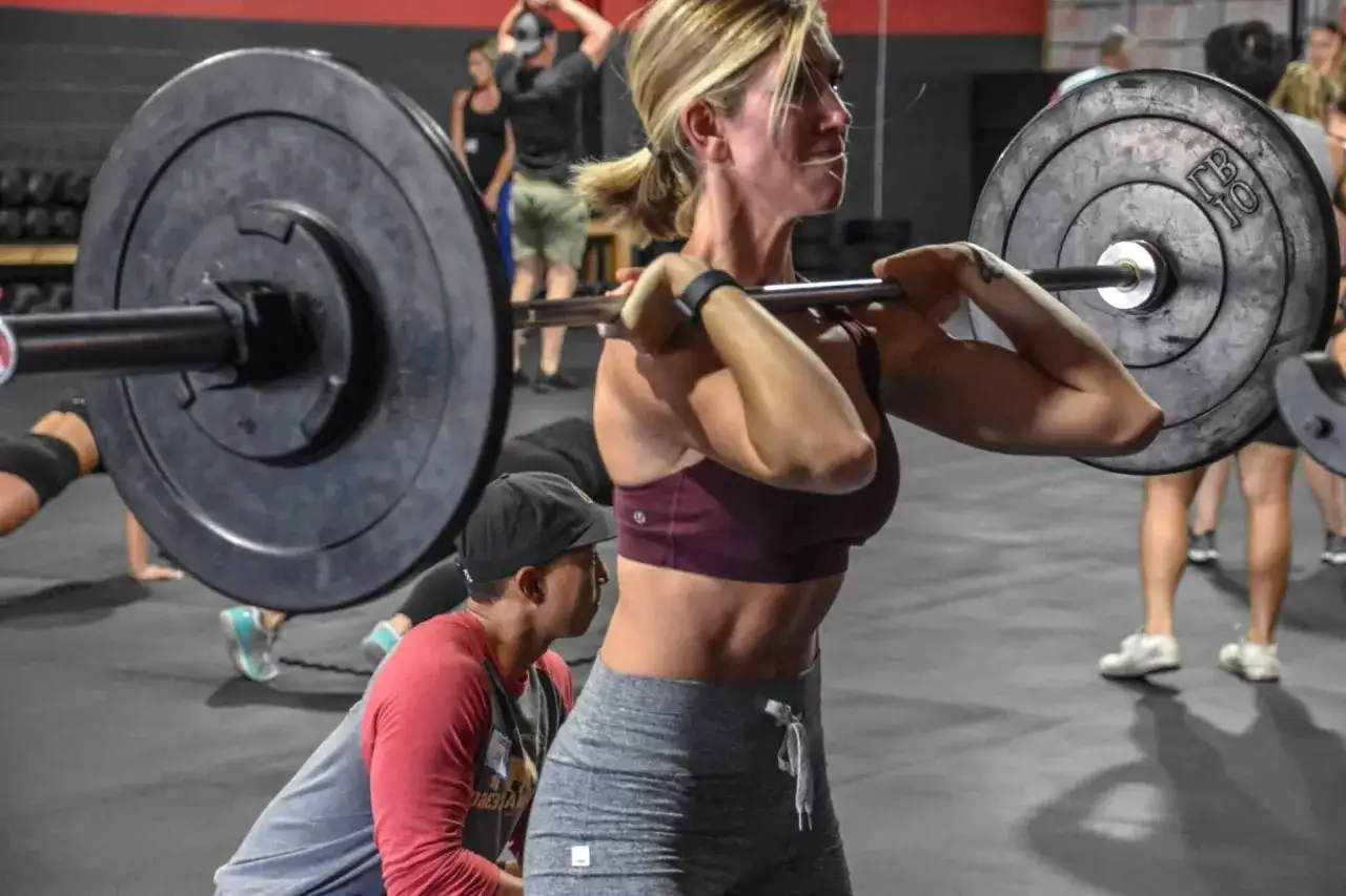 Une femme déterminée commence la musculation, soulevant une barre chargée. Un homme observe, prêt à aider.