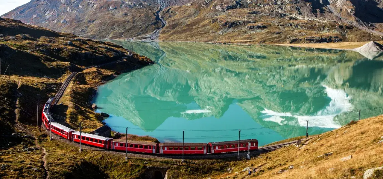 A red train winds its way along a turquoise lake, reflecting the rugged mountains. The journey from Chur to Tirano offers breathtaking views.
