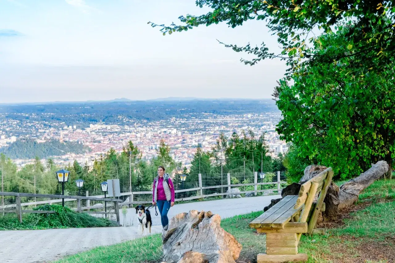 Frau mit Hund spaziert auf einem Weg mit Blick auf Graz. Ein perfekter Ort für graz geheimtipps und Ausblicke.
