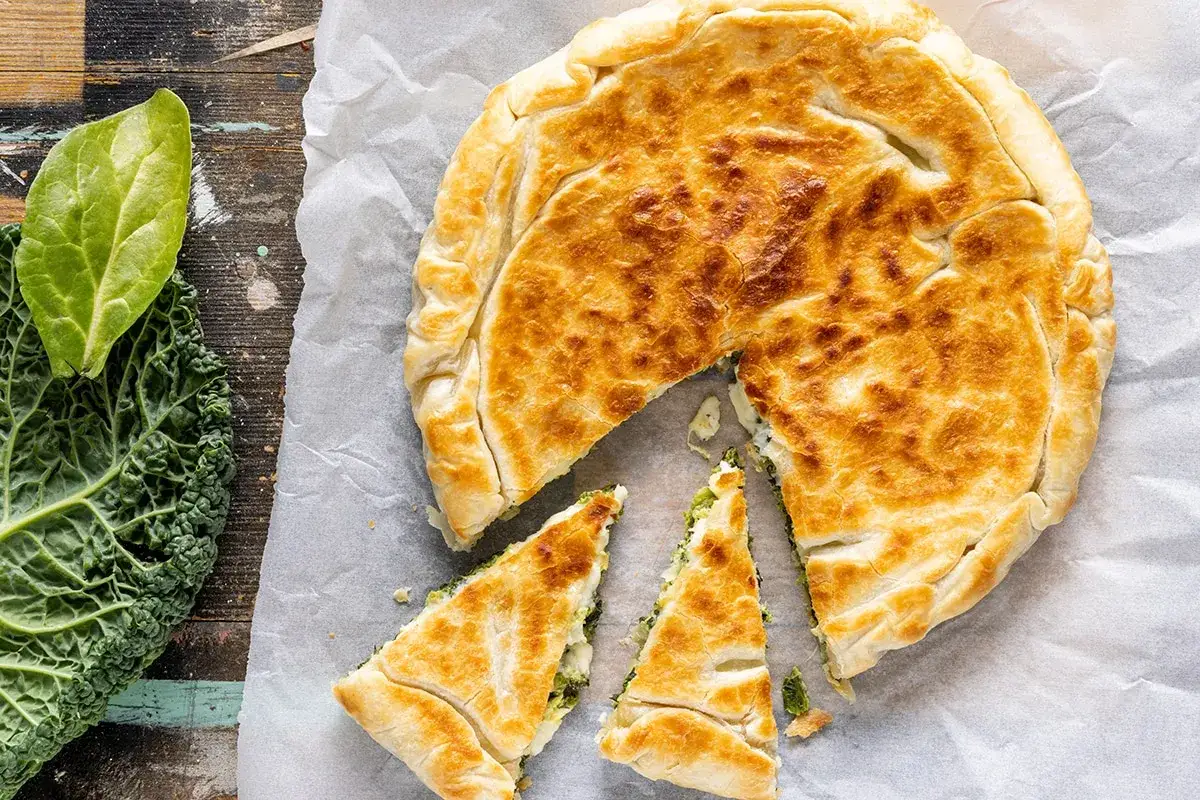 Torta salata con pasta sfoglia dorata, ripiena di verdure. Accanto, foglie di cavolo e spinaci freschi.