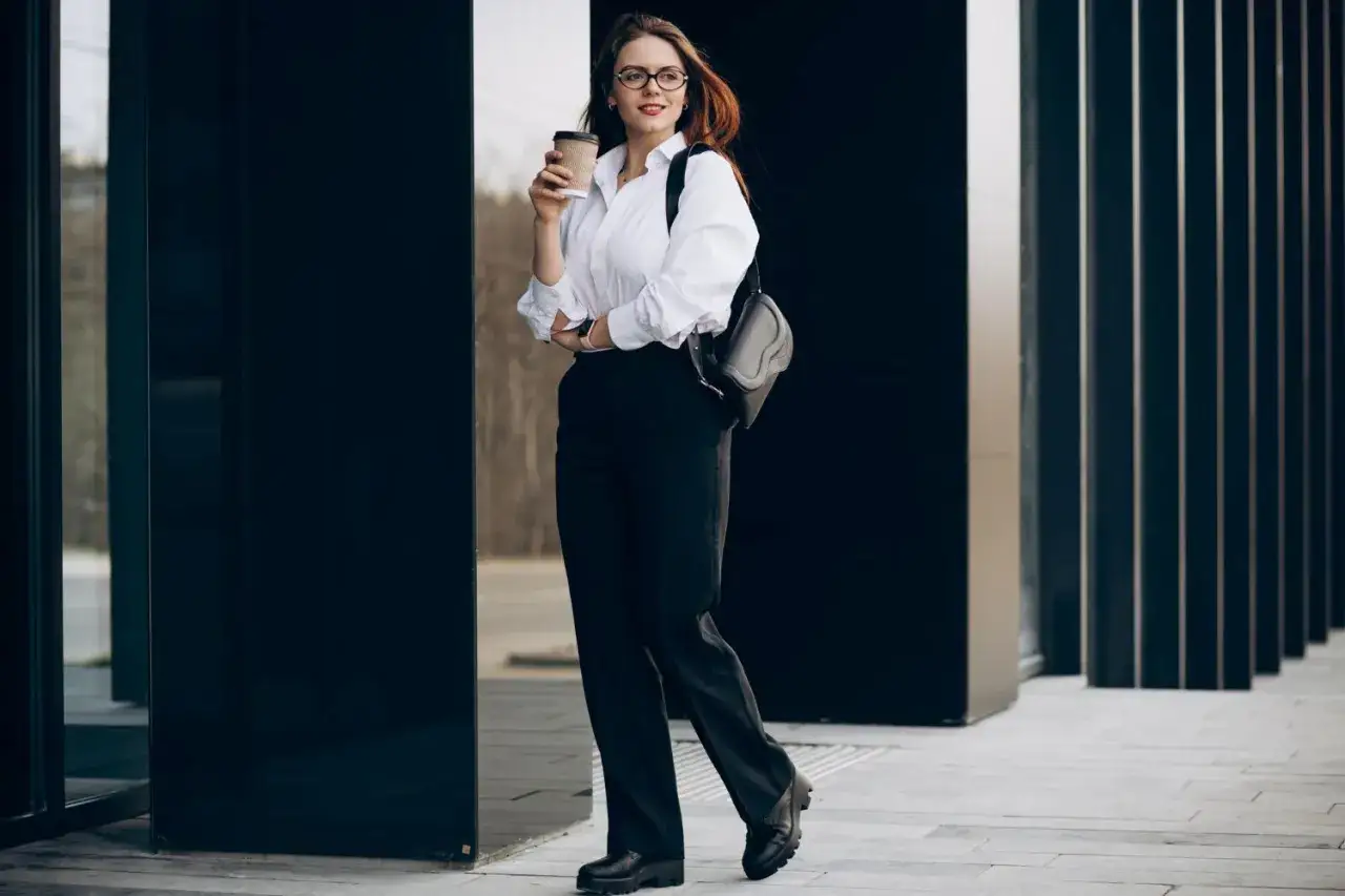 Elegancki strój na maturę damski: biała koszula, czarne spodnie, buty na platformie i torebka. Kobieta trzyma kubek kawy.