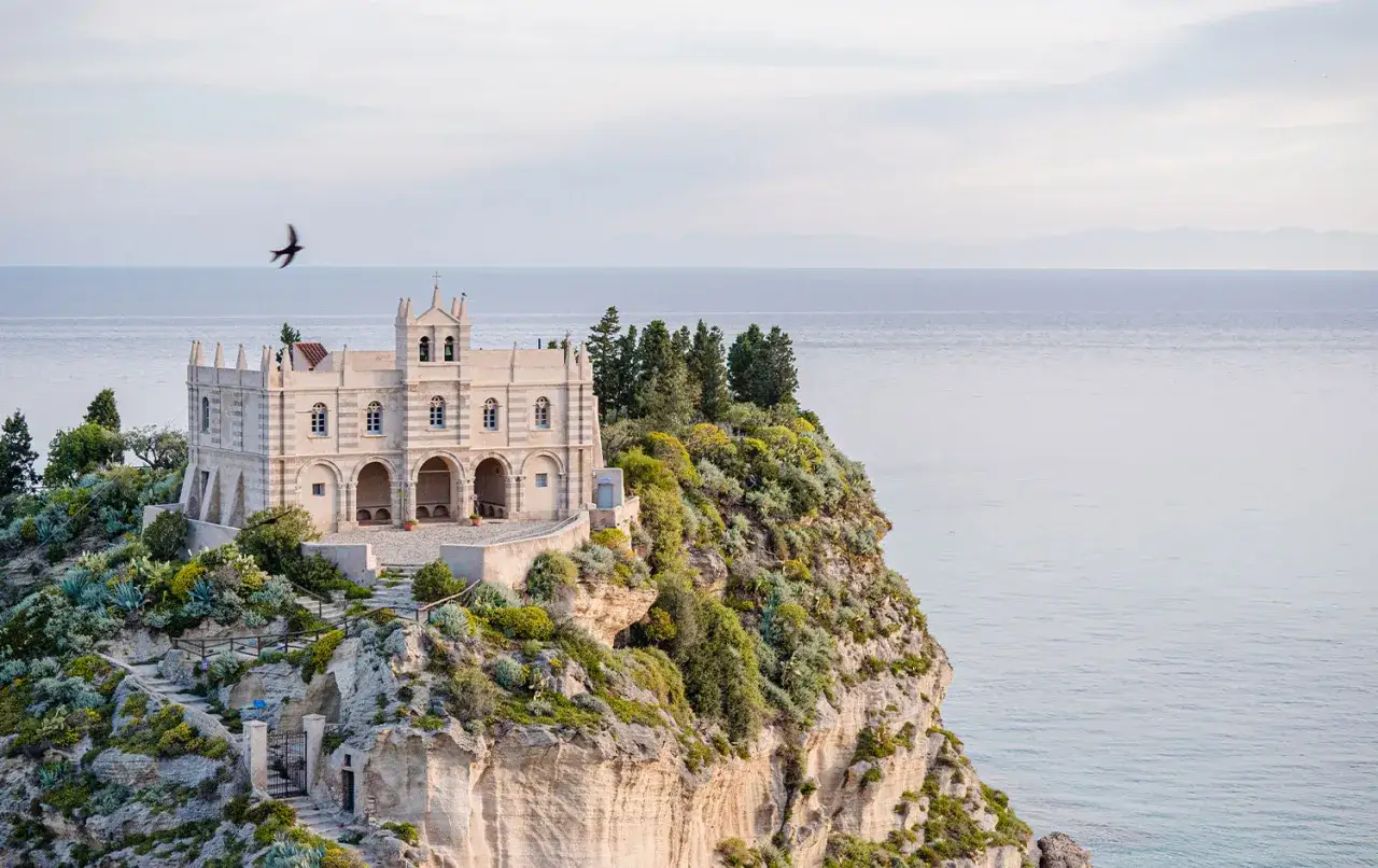 La chiesa di Tropea, un gioiello su una scogliera, domina il mare azzurro. Un gabbiano vola libero nel cielo.