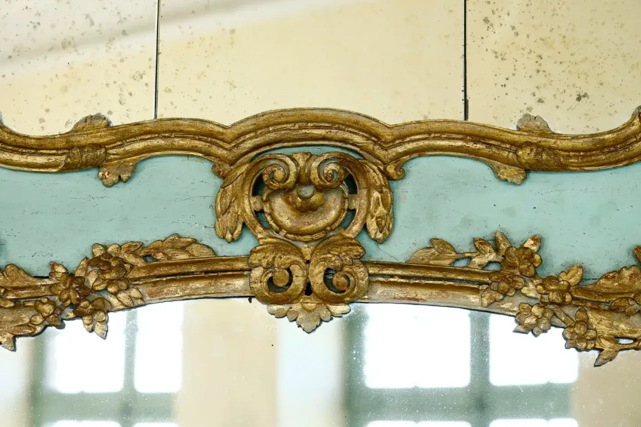Ornement dor&eacute; sculpt&eacute;, typique du style **rococo def.**, orne un miroir ancien au cadre bleu p&acirc;le. Des volutes et des fleurs s'entrem&ecirc;lent.