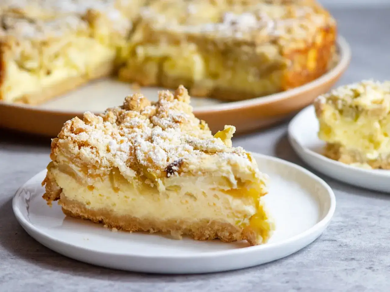 Une part de gâteau mascarpone pommes, saupoudrée de sucre glace, sur une assiette blanche. Le reste du gâteau est visible en arrière-plan.