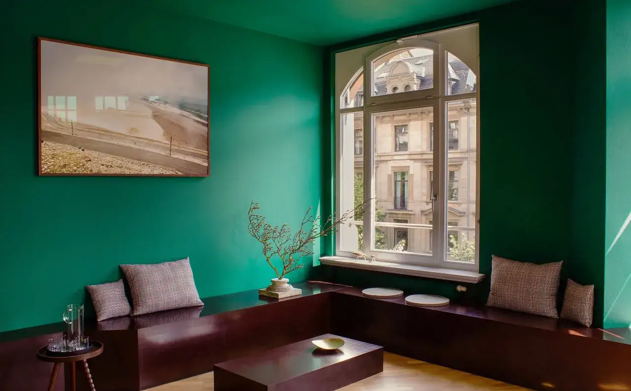 Un soggiorno con pareti verde scuro, un quadro astratto, sedute in legno scuro e cuscini a quadri. I colori da abbinare al verde scuro includono il marrone del legno e le sfumature neutre dei cuscini.