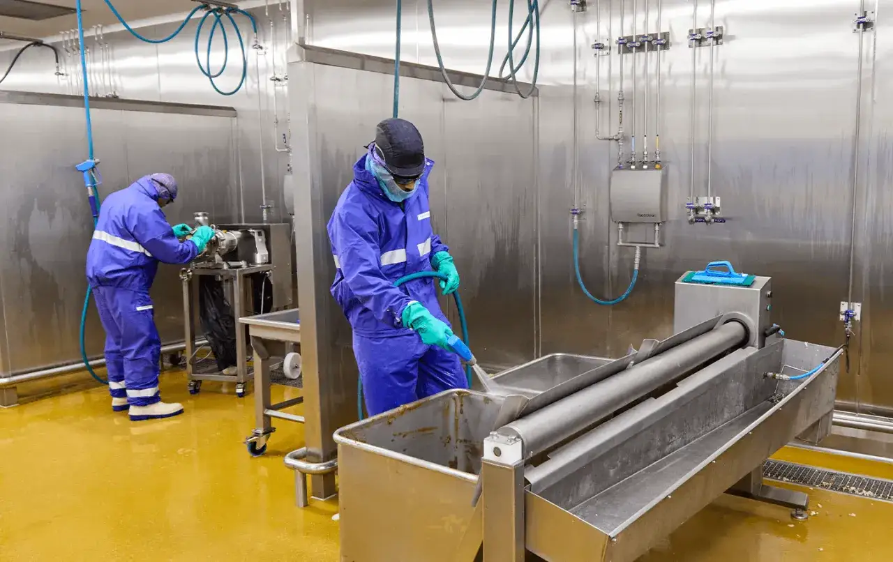 Dos operarios con trajes azules realizan tareas de limpieza en una planta industrial, utilizando mangueras y equipos para mantener los sistemas de limpieza impecables.